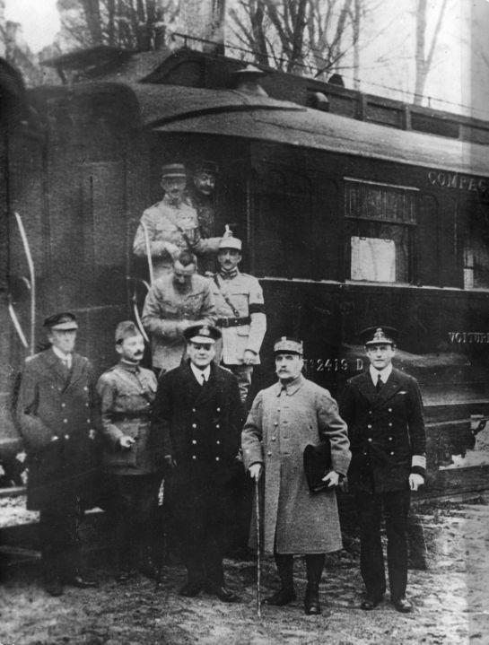 A black-and-white photograph of Allied officers in greatcoats standing in front of a railway carriage in a forest, with more officers on its steps