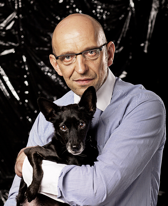 Janusz Chabior in glasses and a striped shirt, holding a small black dog, against a dark background