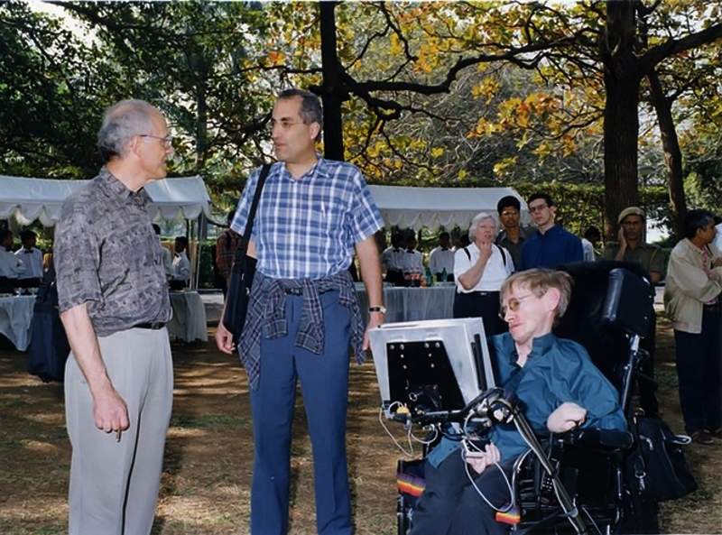 Three men outdoors under trees: an older grey-haired man, a man in a checked shirt, and Stephen Hawking in his wheelchair