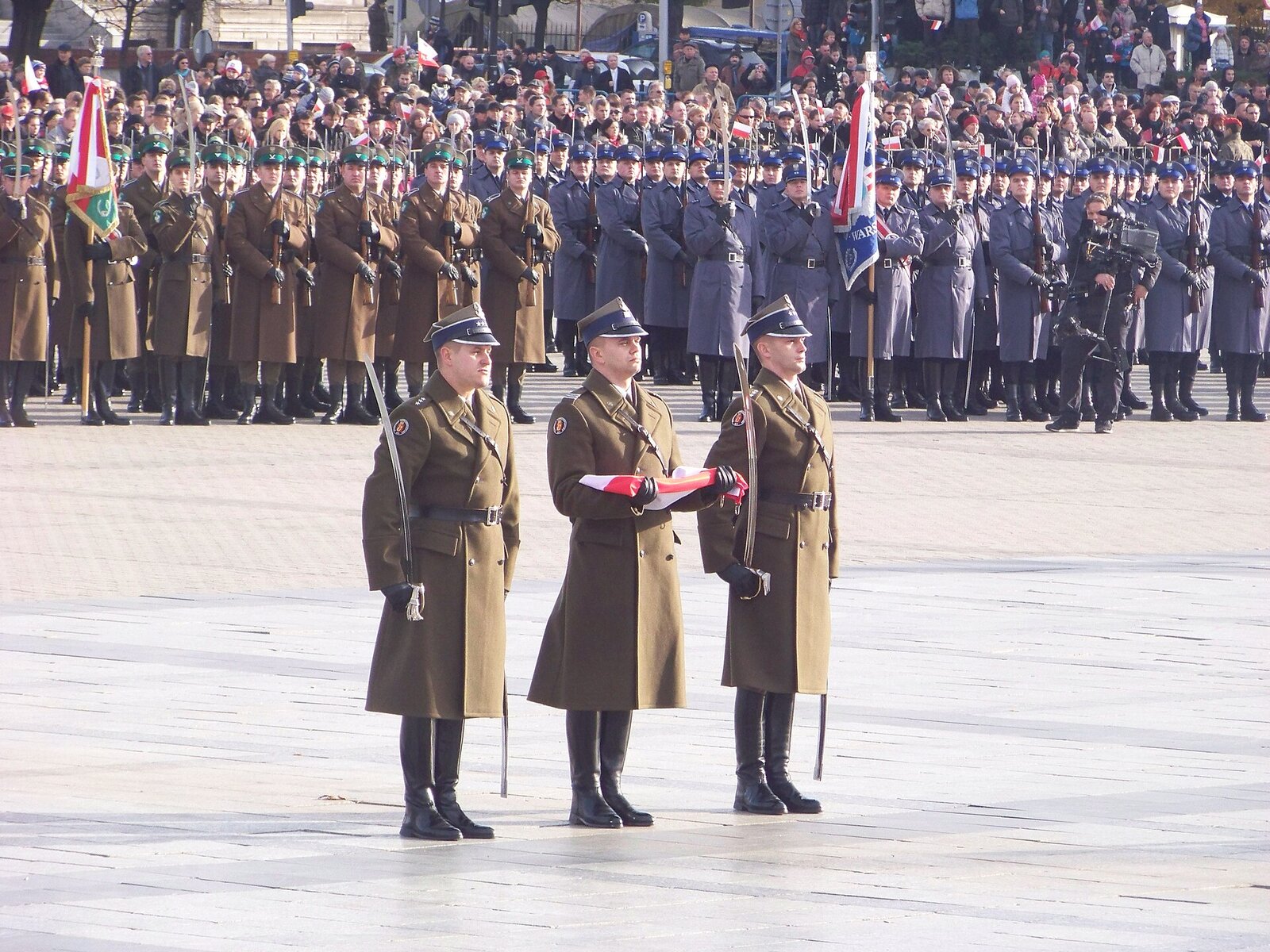 Three Polish soldiers in long olive greatcoats, the one in the middle holding a folded white-and-red flag, with rows of soldiers and a crowd with small flags behind them