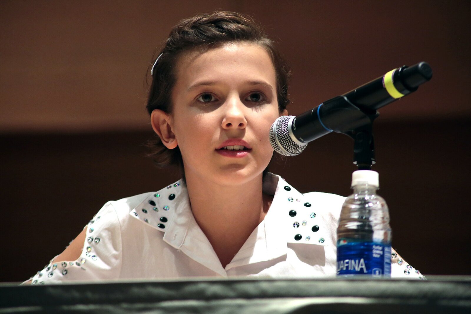 Millie Bobby Brown, a girl with short dark hair in a white studded shirt, speaking into a microphone at a panel table