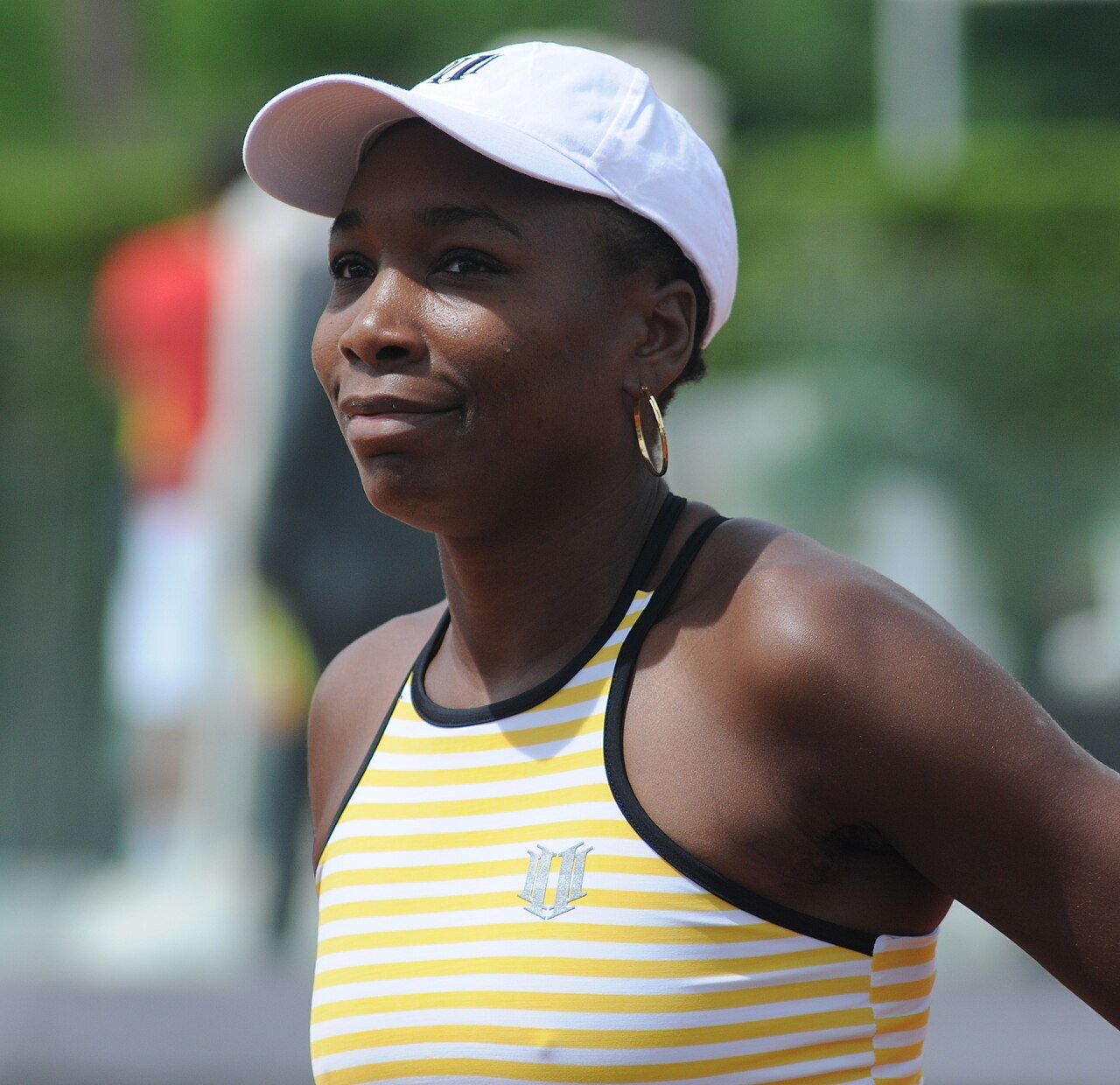 Venus Williams in a white cap and a yellow-and-white striped top; on her chest and on the cap is a logo shaped like a V made of two ones