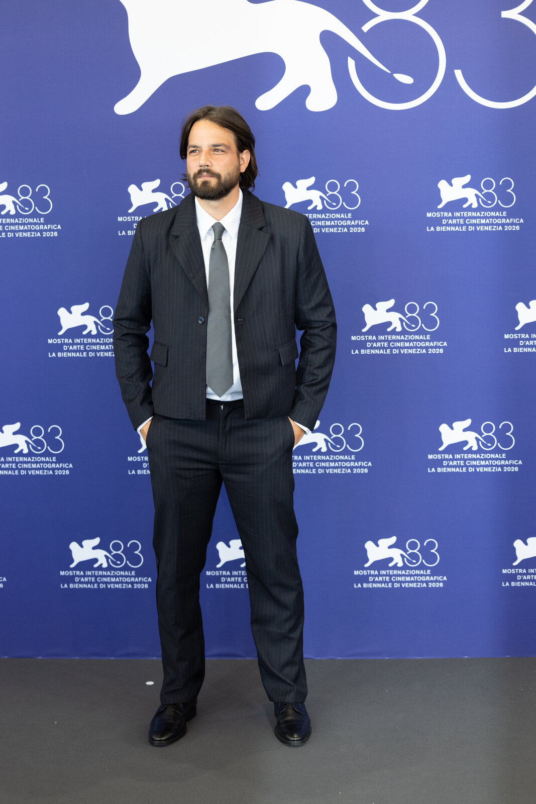 Daniel Zovatto, bearded, in a dark pinstriped suit and grey tie, standing in front of a blue Venice Film Festival backdrop
